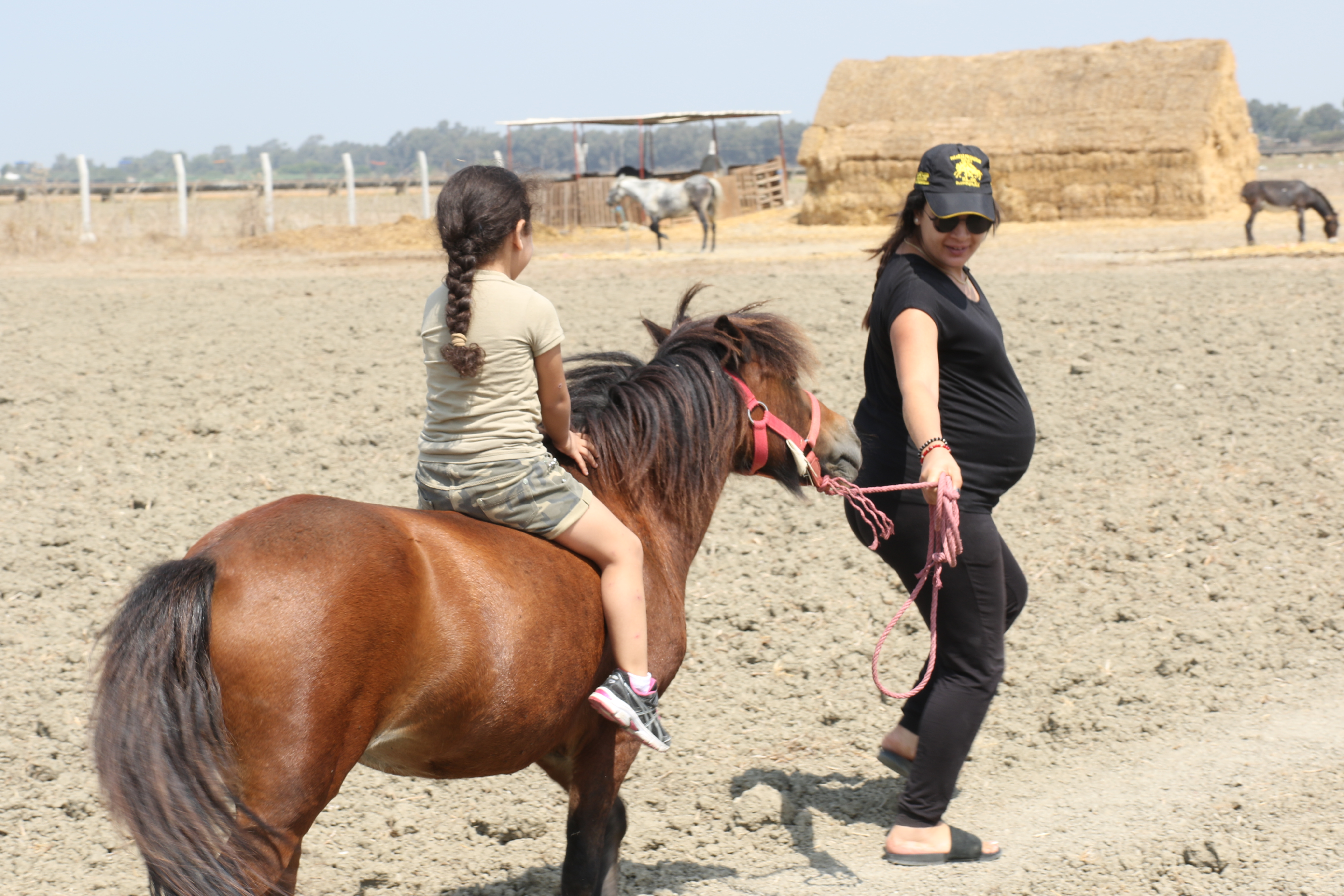 Pregnant mother in jeans and a t-shirt leads a pony that is shorter than she is. Her dauther sit on the pony without a saddle.