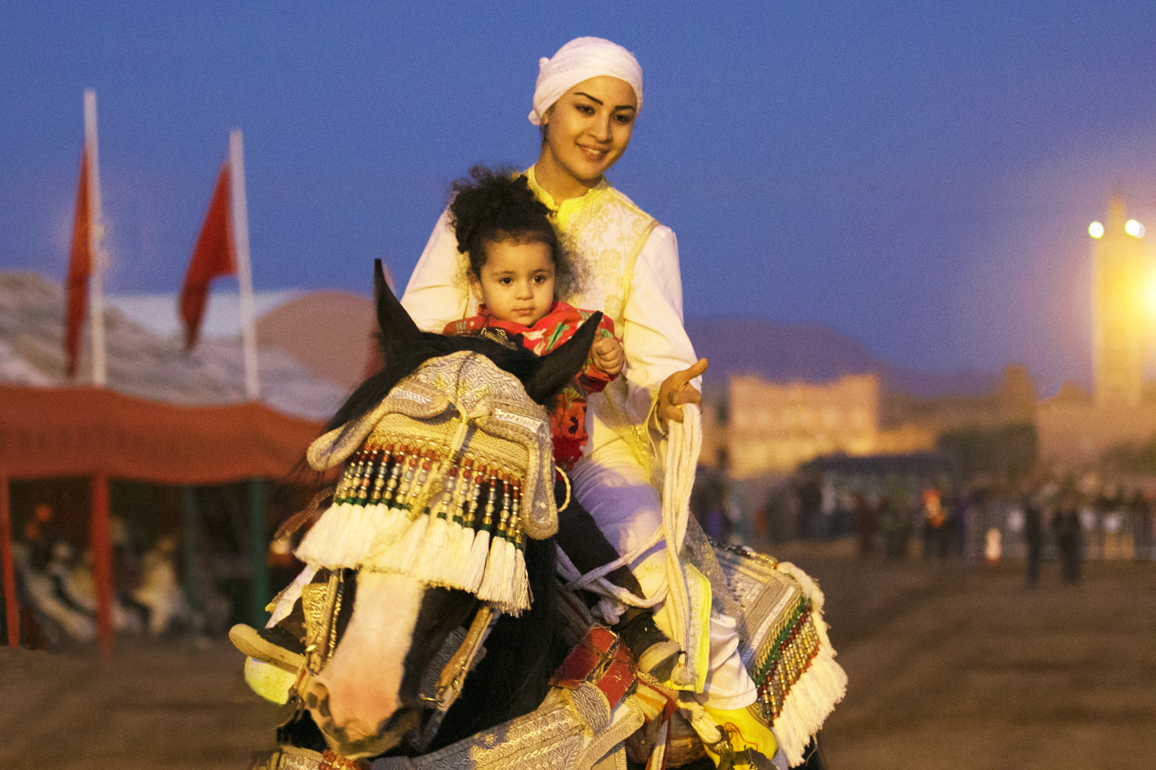 Lilia, no more than three years old, sits in front of Amal atop a horse. Decorative tassels cover the horse's eyes.