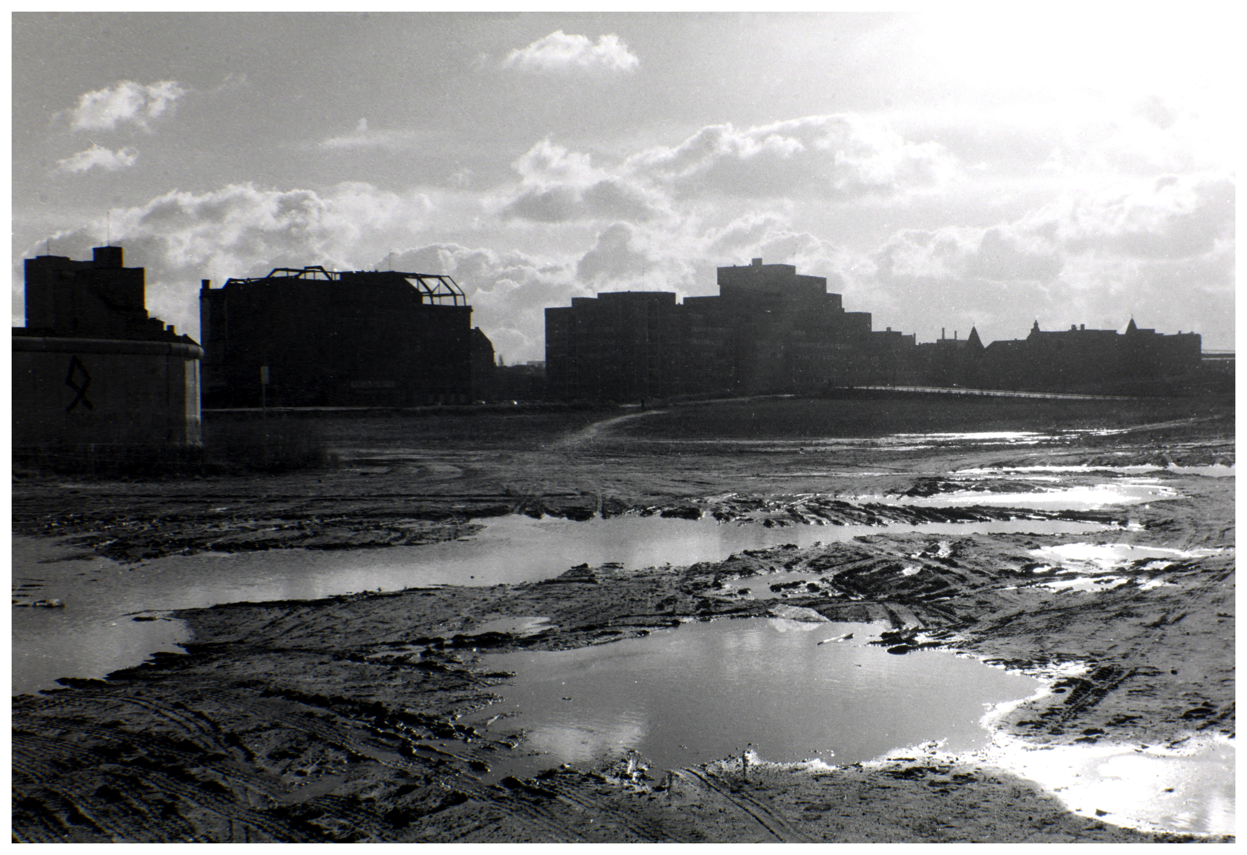 A black and white photo of an open, muddy area and buildings lining the horizon.