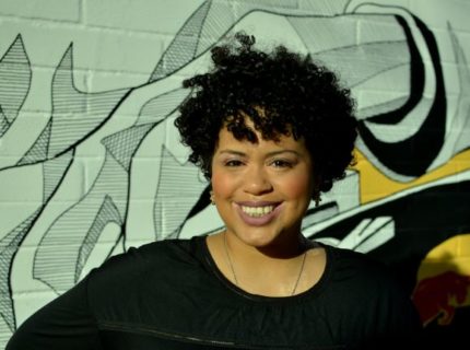 A photograph of Denise Diaz, a Black Latina woman, smiling, wearing a black shirt, and standing in front of a mural