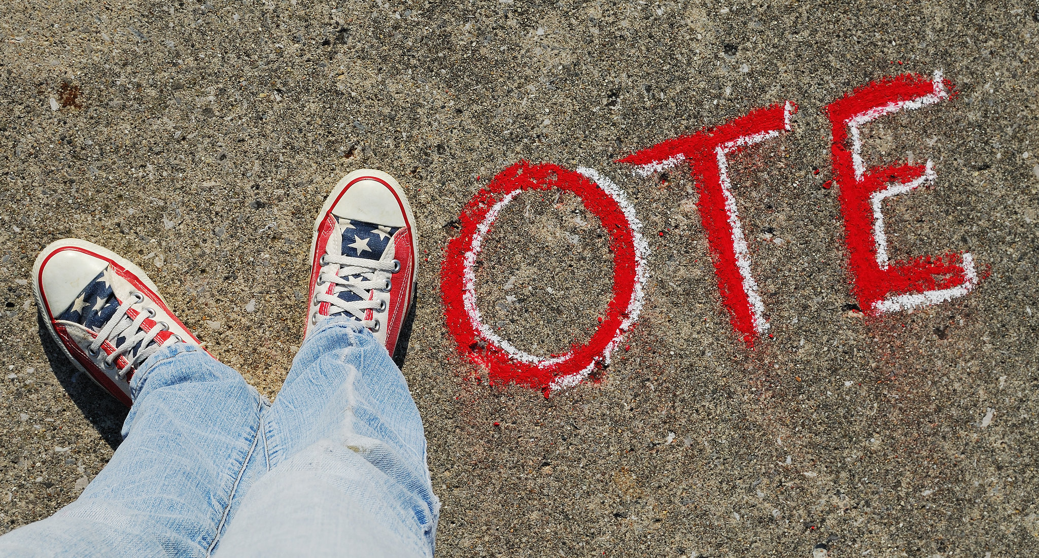 Photograph of two feet in red, white, and blue sneakers compose the letter "V," followed by "ote" in sidewalk chalk.